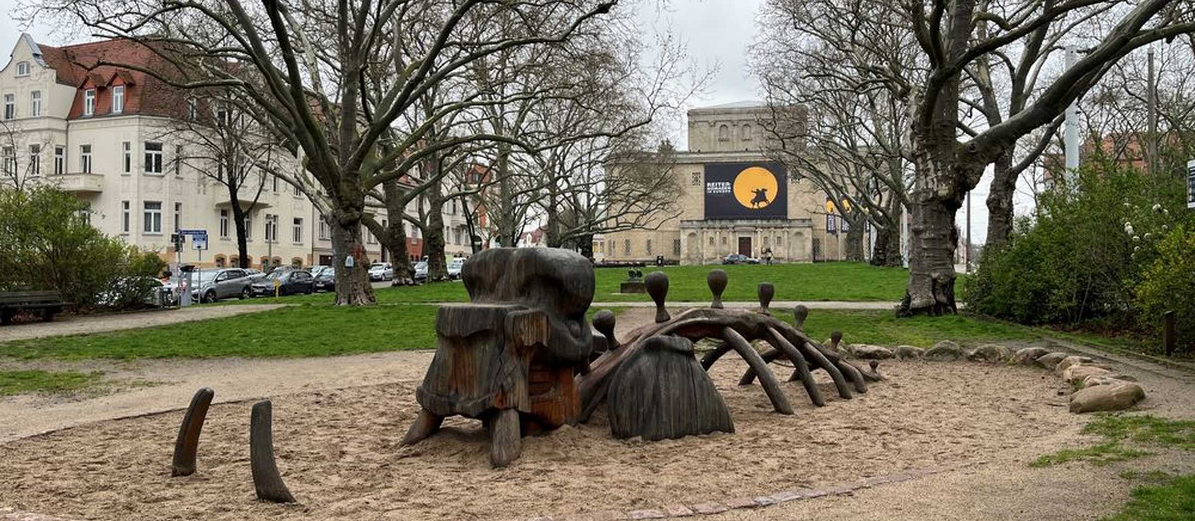 Im Hintergrund die Fassade des Landesmuseums, im Vordergrund das hölzerne Spielgerüst in Form eines Mammuts, teilweise eingegraben im Sand eines Kinderspielplatzes.