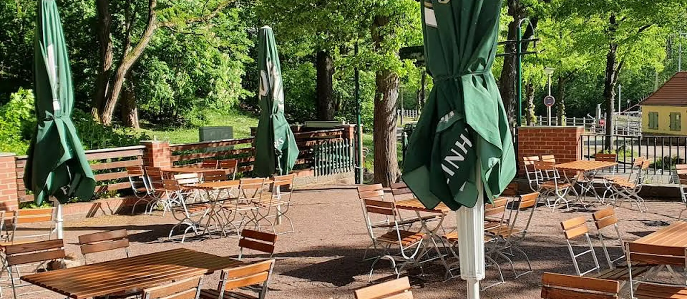 Blick auf den Biergarten des FelsenPavillons mit Sonnenschirmen und Holzstühlen und -tischen in einer grünen Umgebung.