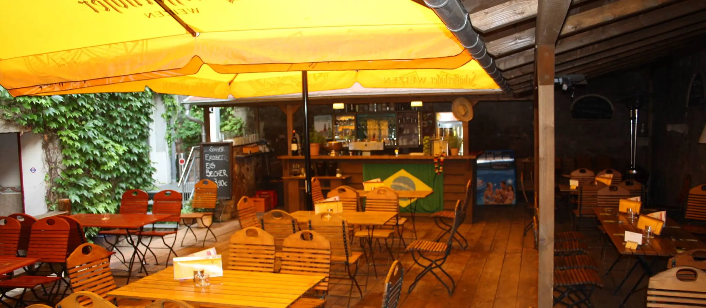 Blick auf die Terrasse des Café Deix mit gelben Sonnenschirmen und Holztischen. Im Hintergrund sieht man eine kleine Bar.