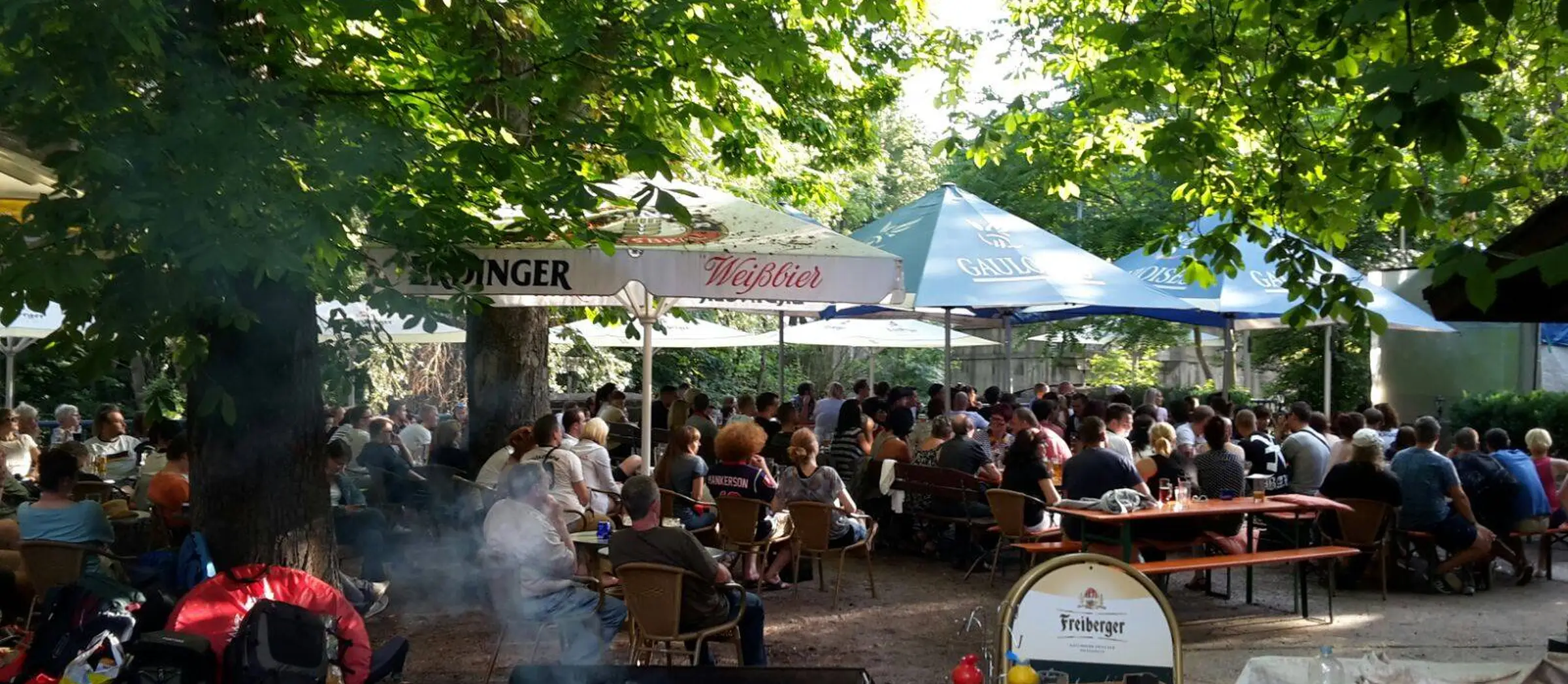 Viele Menschen sitzen im  Freisitz des Fritzengartens unter Sonnenschirmen direkt am Ufer der Saale. Im Vordergrund steht ein Grill.
