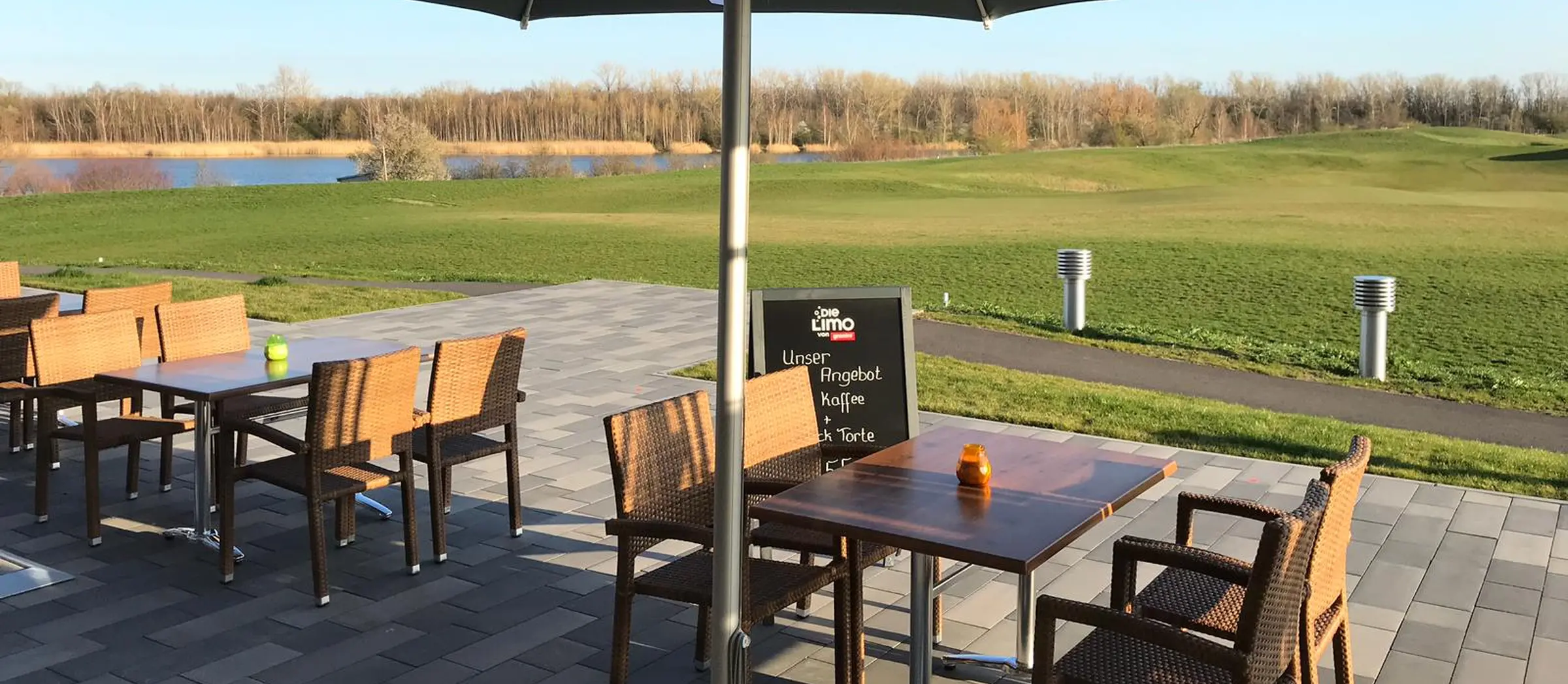 Blick auf die Außenterrasse, im Hintergrund befinden sich ein weiter grüner Rasen und ein See