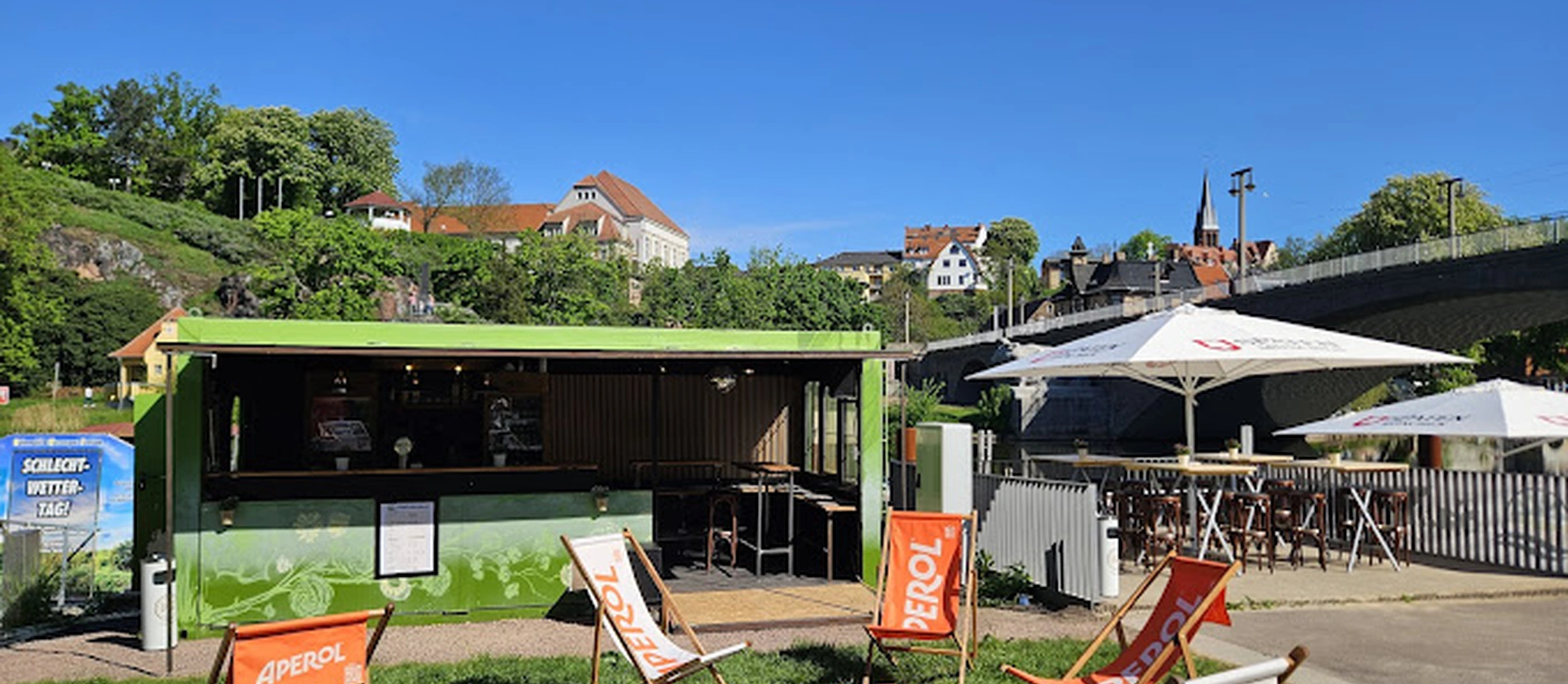 Das Bild zeigt eine Bar mit vereinzelten Liegestühlen auf einer Wiese, direkt an der Saale gelegen. Im Hintergrund sind das Stadtbild von Kröllwitz und die Kröllwitzbrücke zu sehen.