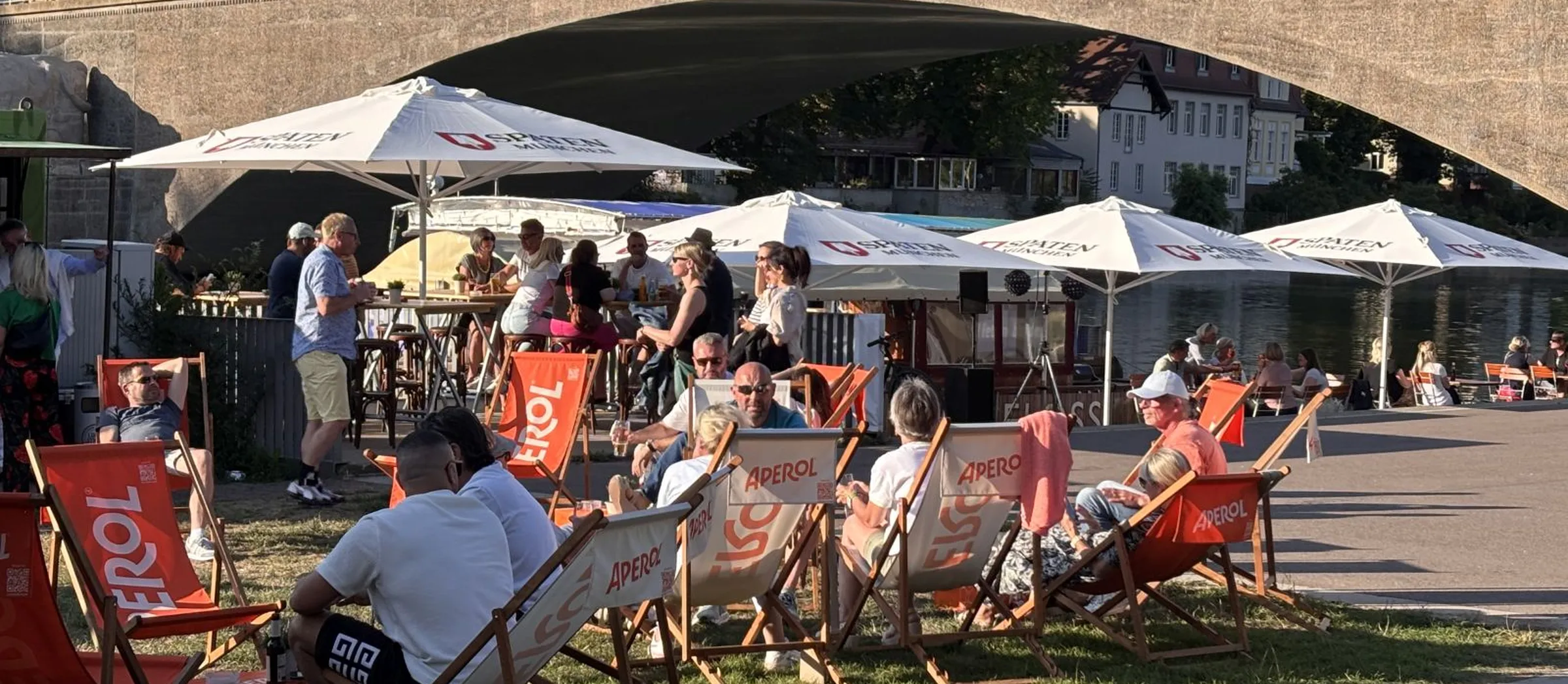 Das Bild zeigt eine sommerliche Stimmung mit chillenden Menschen, direkt an der Saale gelegen. Im Hintergrund sind das Stadtbild von Kröllwitz und die Kröllwitzbrücke zu sehen.