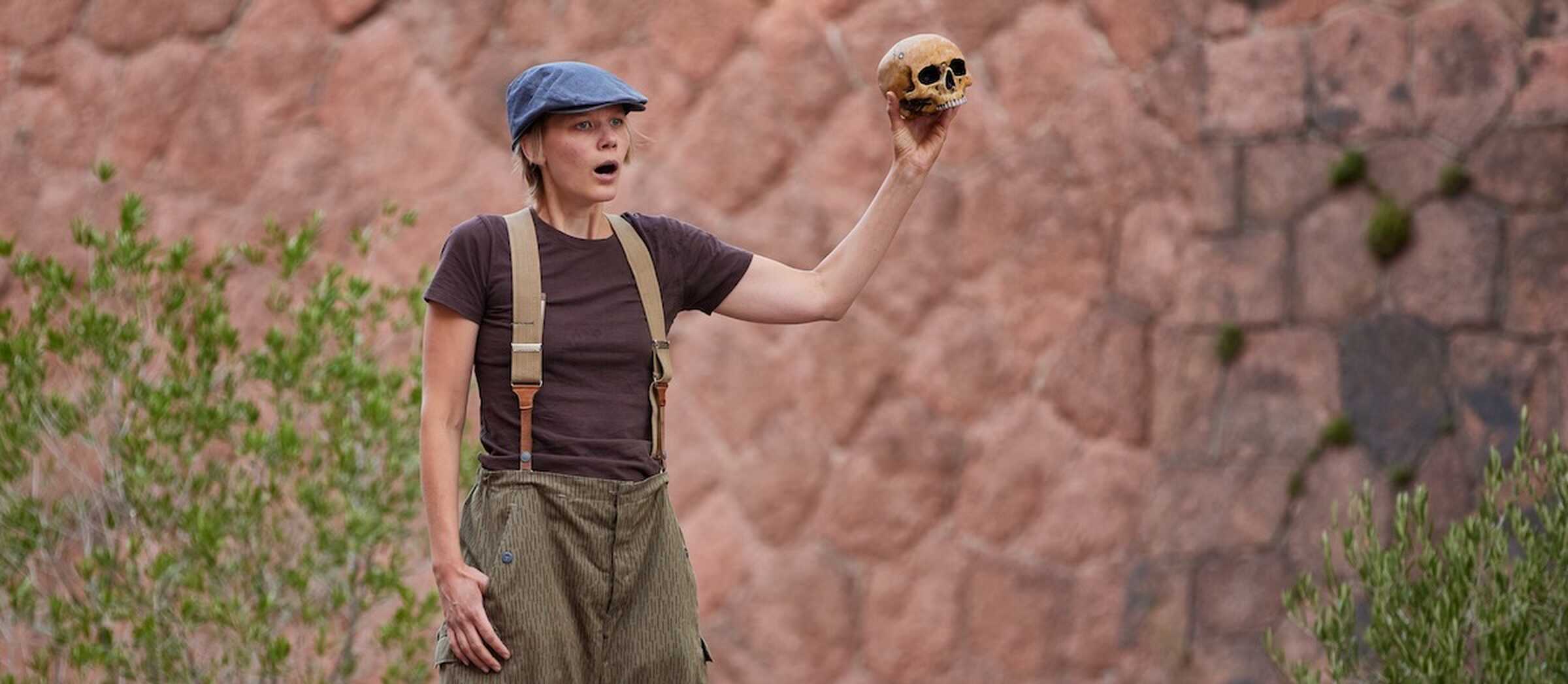 Eine Schauspielerin steht auf einer Open-Air-Bühne und spricht, während sie einen Totenschädel hochhält.
