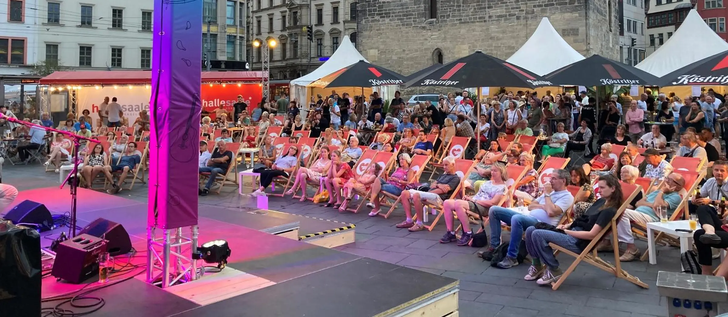Vor einer beleuchteten Bühne in Abendstimmung sitzt das Publikum in Liegestühlen.