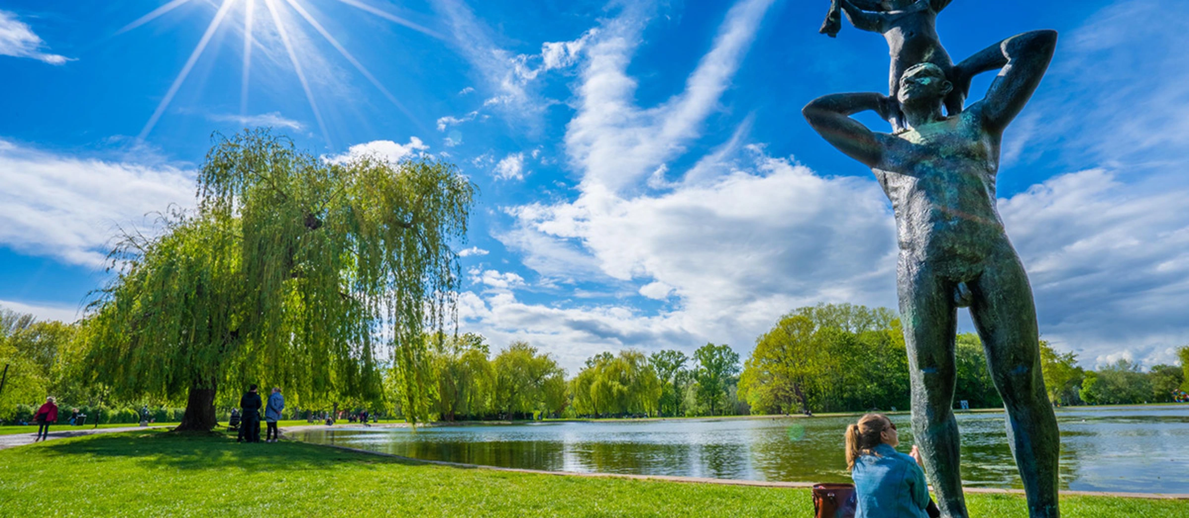 Im Vordergrund sieht man eine Statue, im Hintergrund scheint die Sonne über die Ziegelwiese am Fontäneteich