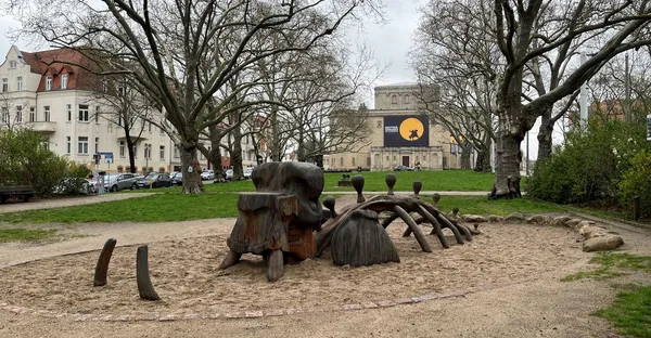 Im Hintergrund die Fassade des Landesmuseums, im Vordergrund das hölzerne Spielgerüst in Form eines Mammuts, teilweise eingegraben im Sand eines Kinderspielplatzes.