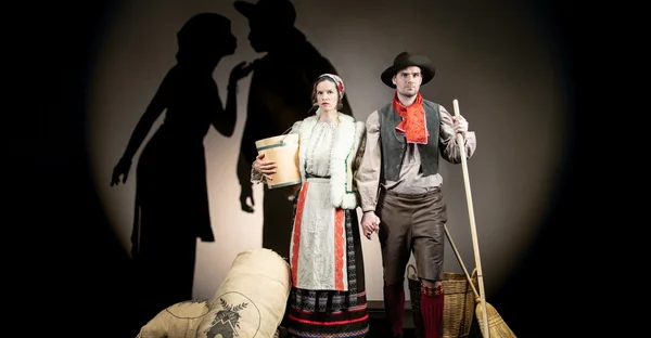 Sybille Kreß und Marian Kindermann stehen in traditioneller Robe zwischen Besen und Getreidesäcken. Im Hintergrund sind Schatten von zwei Personen, die sich küssen. 