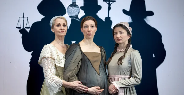 Elke Richter hält den schwangeren Bauch von Nicoline Schubert, während diese Kinga Schmidts Hand hält. Im Hintergrund sind drei Personen mit Hüten, welche Waage, Reagenzglas und Kruzifix in den Händen halten. 