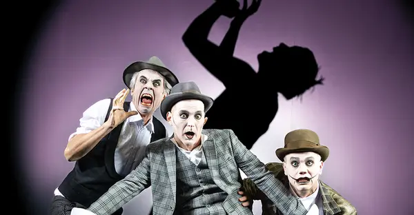 Enrico Petters, Till Schmidt und Florian Krannich tragen Anzüge und sind weiß geschminkt. Sie gucken verängstigt und erschrocken. Im Hintergrund ist ein Schatten einer Person, die singt.