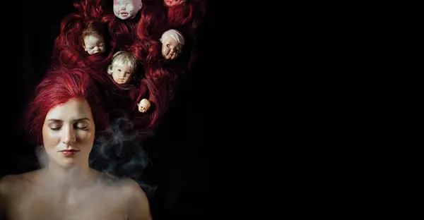 Eine Person liegt mit geschlossen Augen auf einem schwarzen Untergrund. Auf ihren langen, gewellten, roten Haaren liegen sieben Puppenköpfe verteilt. 