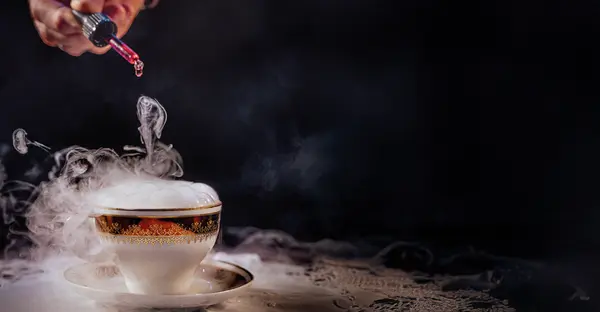 Es qualmt aus einer Tasse, welche auf einem Spitzendeckchen steht. Es wird eine rote Flüssigkeit durch eine Pipette hineingetropft. 