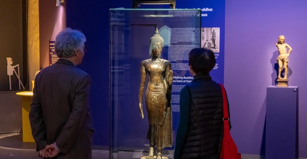 Ansicht der Ausstellung „Fragmente eines Lebens. Die Sammlung Hans-Hasso von Veltheims“ im Kunstmuseum Moritzburg Halle (Saale). Zwei Personen betrachten die Bronze-Plastik einer schreitenden Buddha-Figur. 