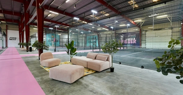 Im Eingangsbereich der Padelhalle befinden sich ein helles Sofa und Pflanzen. Pinke Flure verbinden die Padel-Felder.