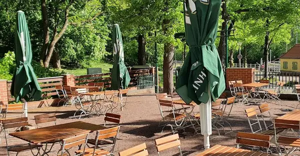 Blick auf den Biergarten des FelsenPavillons mit Sonnenschirmen und Holzstühlen und -tischen in einer grünen Umgebung.