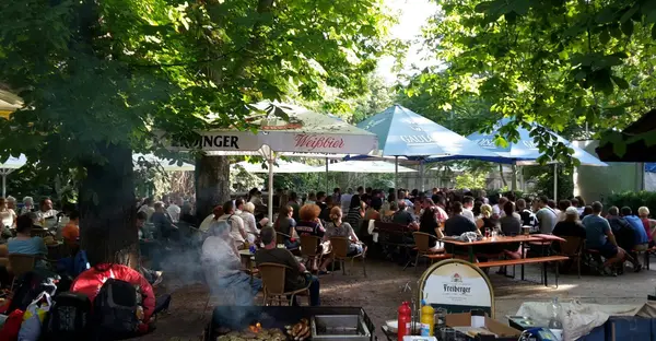 Viele Menschen sitzen im  Freisitz des Fritzengartens unter Sonnenschirmen direkt am Ufer der Saale. Im Vordergrund steht ein Grill.