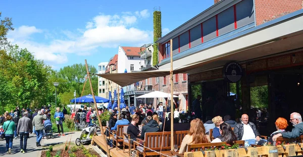Die Außenterrasse des Hafenmeister und Docks ist zu sehen: Viele Menschen sitzen draußen im sommerlichen Ambiente, Bäume sind im Hintergrund und Fußgänger*innen spazieren vorbei.