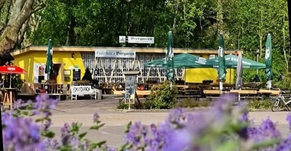 Der Biergarten mit seiner gelben Fassade ist im Hintergrund zu sehen. Im Vordergrund blühen lila Blüten.