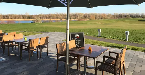 Blick auf die Außenterrasse, im Hintergrund befinden sich ein weiter grüner Rasen und ein See