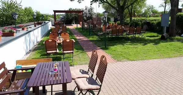 Blick auf die Terrasse der Gaststätte: Holzstühle und -tische stehen verteilt in einer gepflegten Gartenanlage.