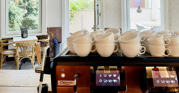 Kaffeetassen stapeln sich auf einer Kaffeemaschine, im Hintergrund ist das Kaffeehaus von innenzu sehen.