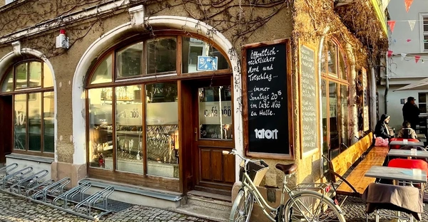 Außenansicht des Café Nöös: Vor der Fassade des Cafés steht ein Fahrrad an die Wand gelehnt, eine Tafel ist mit dem Tagesmenü beschriftet und auf der Terrasse sitzen Gäste.