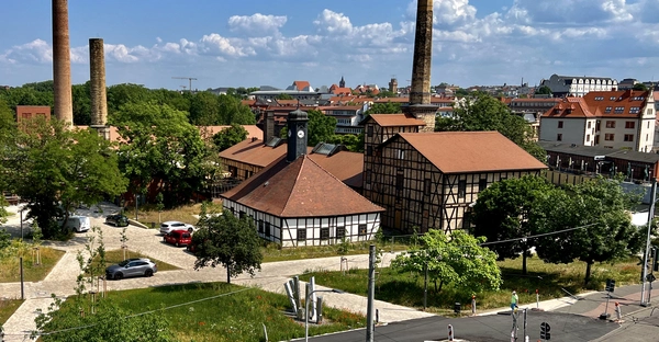 Fachwerkhäuser umgeben von einem Park mit Parkplatz, im Hintergrund Häuser der Stadt