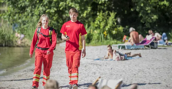 Zwei junge Menschen in roter Bekleidung der DLRG laufen einen Strand eines Sees entlang.