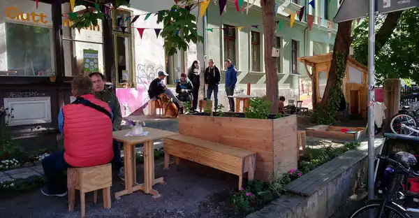 Verschiedene Personen sitzen auf Holzbänken einer Außenterrasse, die mit Girlanden geschmückt ist, oder unterhalten sich im Stehen.