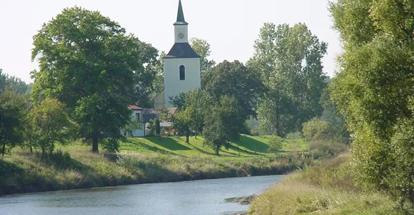 Zu sehen ist eine kleine Kapelle am Fluss gelegen, umgeben von grüner Landschaft.