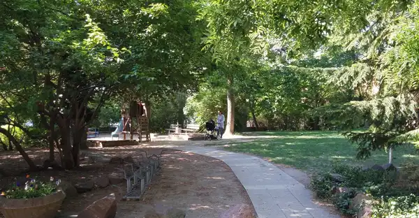 Zu sehen ist eine schattige Parkanlage mit Spazierwegen und einem Spielplatz im Hintergrund.