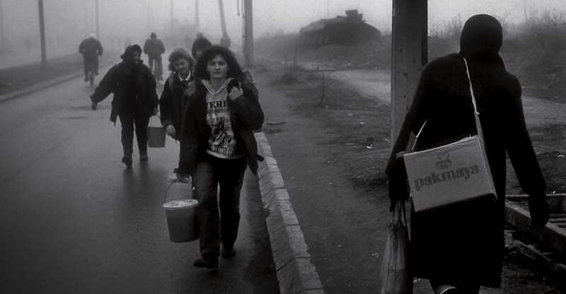 Blick in eine Straße, Menschen tragen Gepäck. Im Hintergrund liegt ein Panzer. Fotografie in schwarz/weiß. Sarajevo-1993 © Knut Mueller