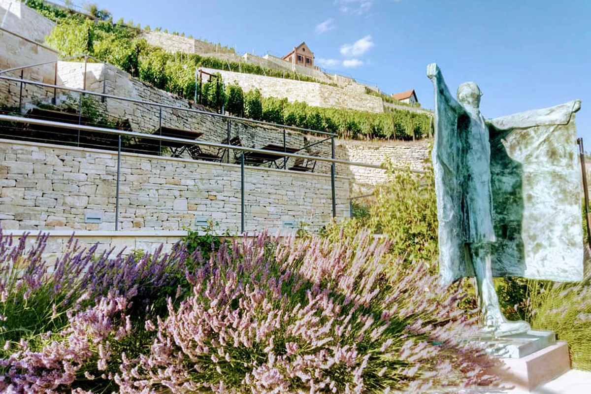 Im Vordergrund ist eine Statue und blühender Flieder zu sehen, im Hintergrund ein Weinberg mit Weinterrassen.