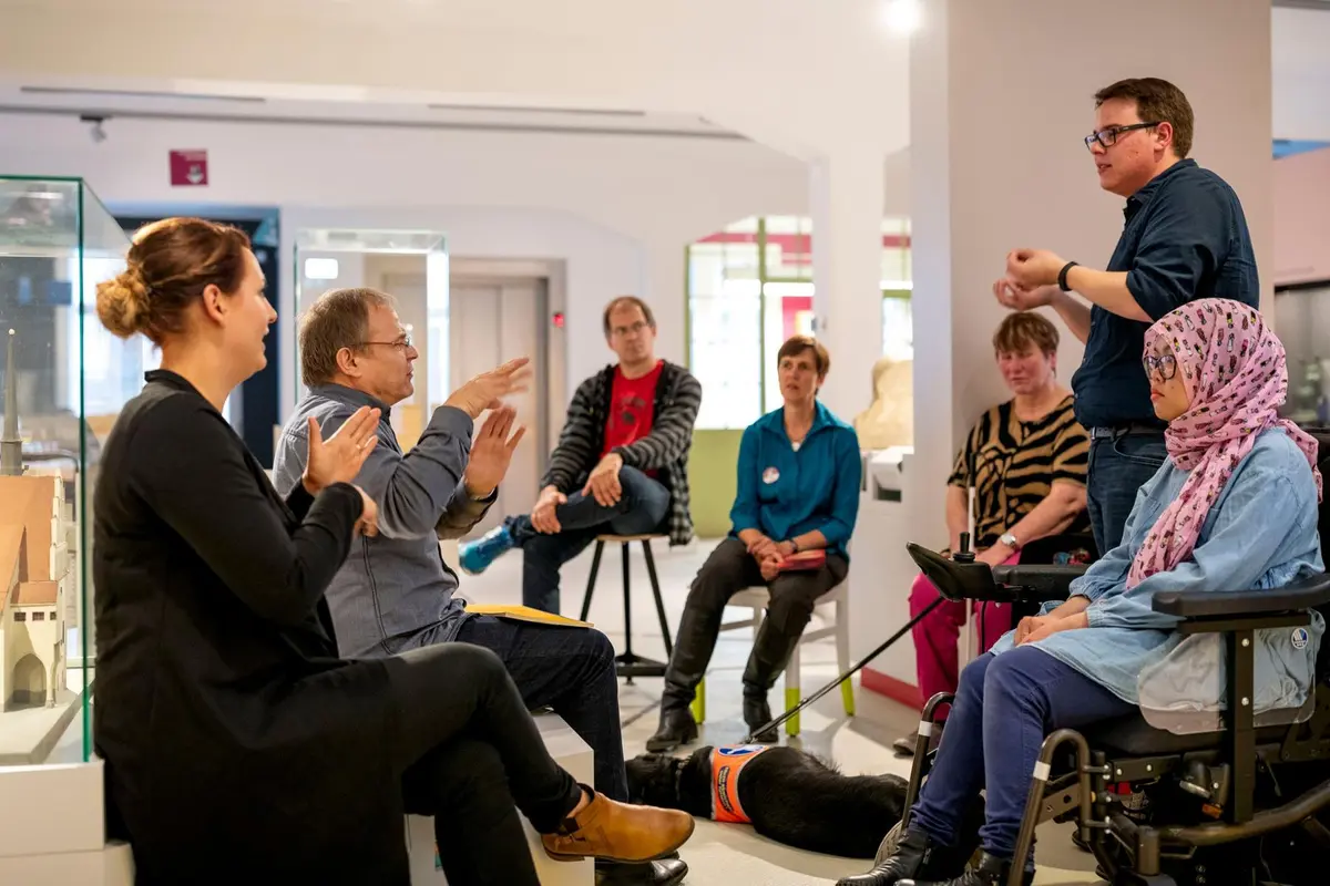 Mehrere Personen sitzen auf Stühlen, es wird in Gebärdensprache gesprochen.