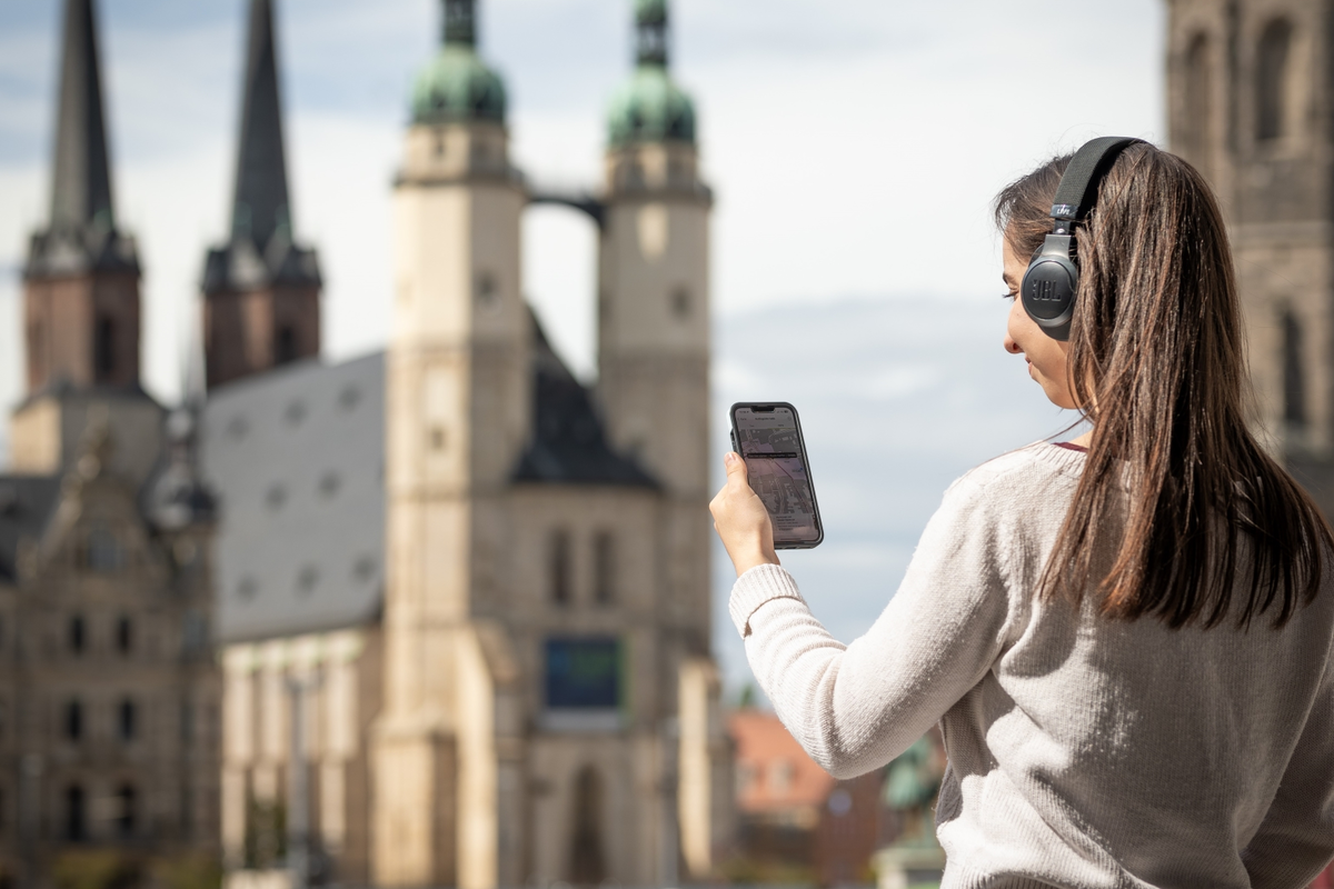 Ein junge Frau steht auf dem Marktplatz in Halle und hält ihr Handy in der Hand. Sie trägt Kopfhörer und im Hintergrund sind die Hausmannstürme zu sehen.