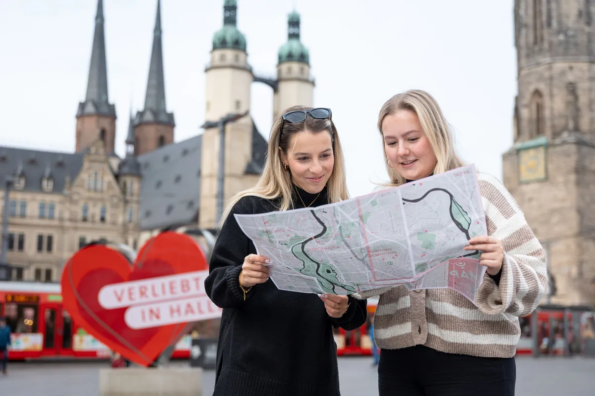 Zwei junge Frauen stehen auf dem Marktplatz in Halle und schauen in einen Stadtplan. Im Hintergrund sind die Hausmannstürme und das "Verliebt in Halle" Herz zu sehen.
