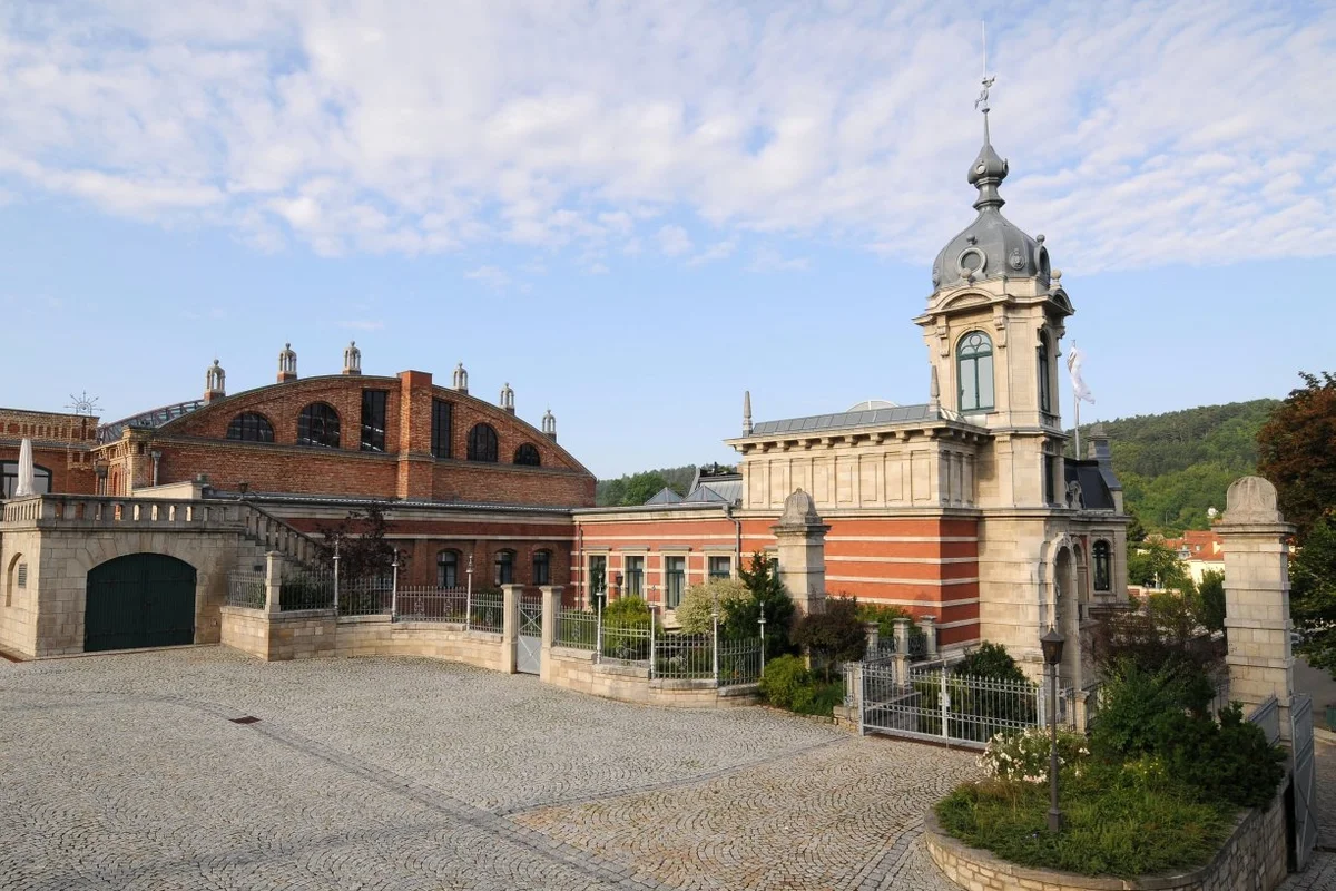 Zu sehen ist ein kleines schloss-artiges Gebäude mit einem Turm. Es hat eine rote Fassade. Im Hintergrund ist ein bewaldeter Hügel zu sehen..
