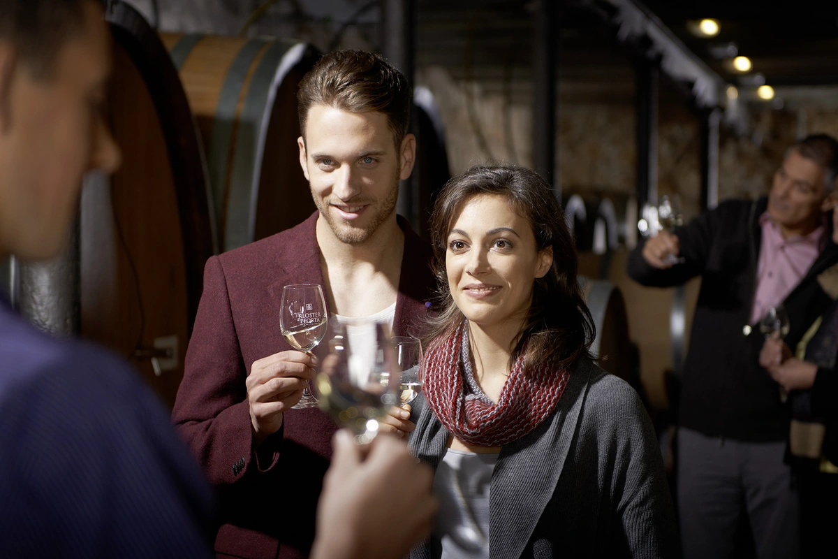 Eine Frau und ein Mann stehen in einem Weinkeller und halten beide ein Glas Wein in der Hand. 